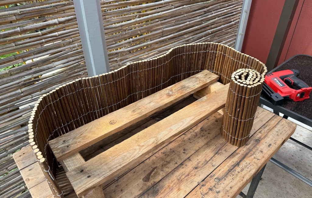Eine kleine Holzpalette, die teilweise mit einer Beizband-Weidenmatte verkleidet ist, steht auf einem Arbeitstisch neben einem roten Einhell Akku-Nagler.