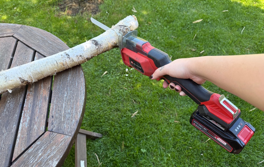 Eine Hand sägt mit einer rot-schwarzen Einhell Akku-Universalsäge einen Birkenstamm durch, der auf einem runden Holztisch liegt.