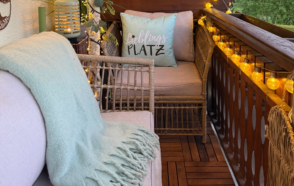 Gemütlich beleuchtete Balkon-Lounge am Abend mit einer eingeschalteten Lichterkette am Holzgeländer und einer hellgrünen Kuscheldecke.