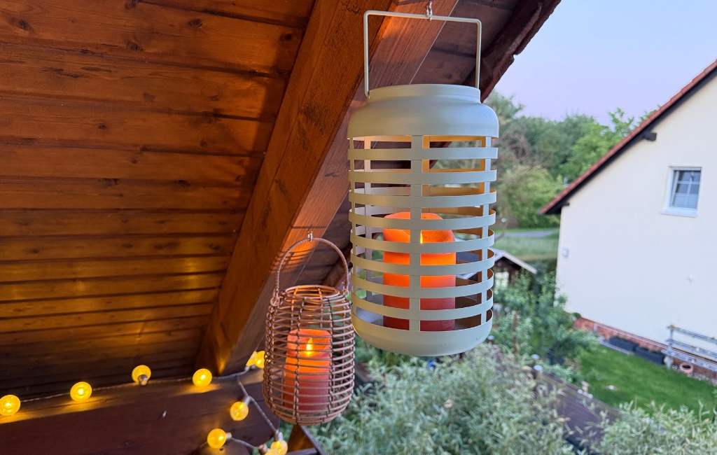 Zwei hängende Windlichter aus Metall und Geflecht mit brennenden LED-Kerzen unter einem hölzernen Balkondach in der Dämmerung.