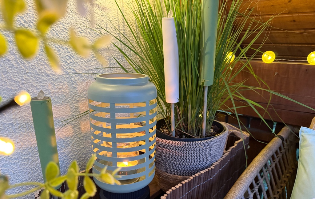 Ein hellgrünes, gemustertes Metall-Windlicht mit brennender Kerze steht neben einer Topfpflanze auf dem Balkon.