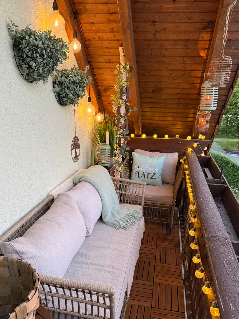 Eine stilvoll eingerichtete Balkon-Lounge am Abend, beleuchtet durch warme Lichterketten, Laternen und Glühbirnen.