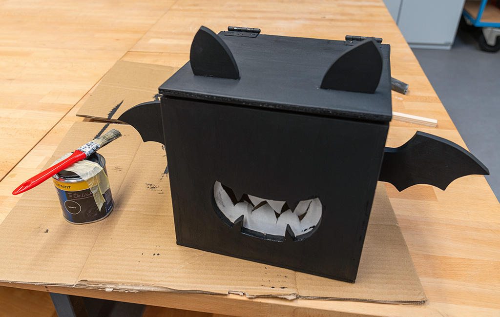 Fertige schwarze Fledermauskiste aus Holz mit Ohren, Flügeln und einem weißen Ausschnitt in Form eines gezackten Mauls, daneben eine Farbdose und ein Pinsel.