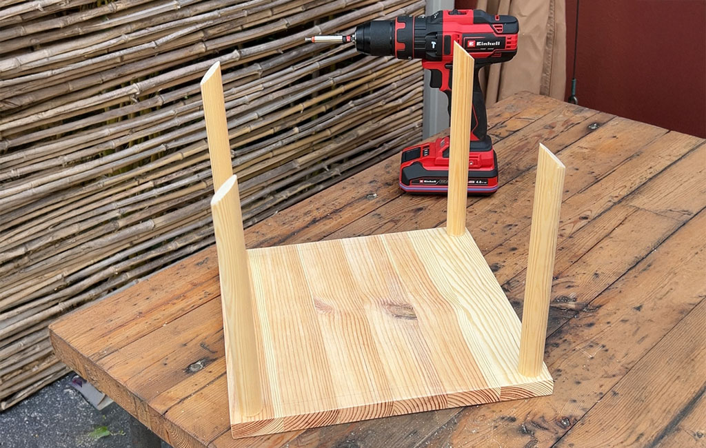 A base plate with four vertical wooden rods and an Einhell cordless drill on a wooden table.