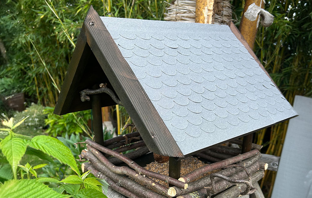 Bird feeder made of branches, wood, and grey roof placed on a tree stump in the garden.