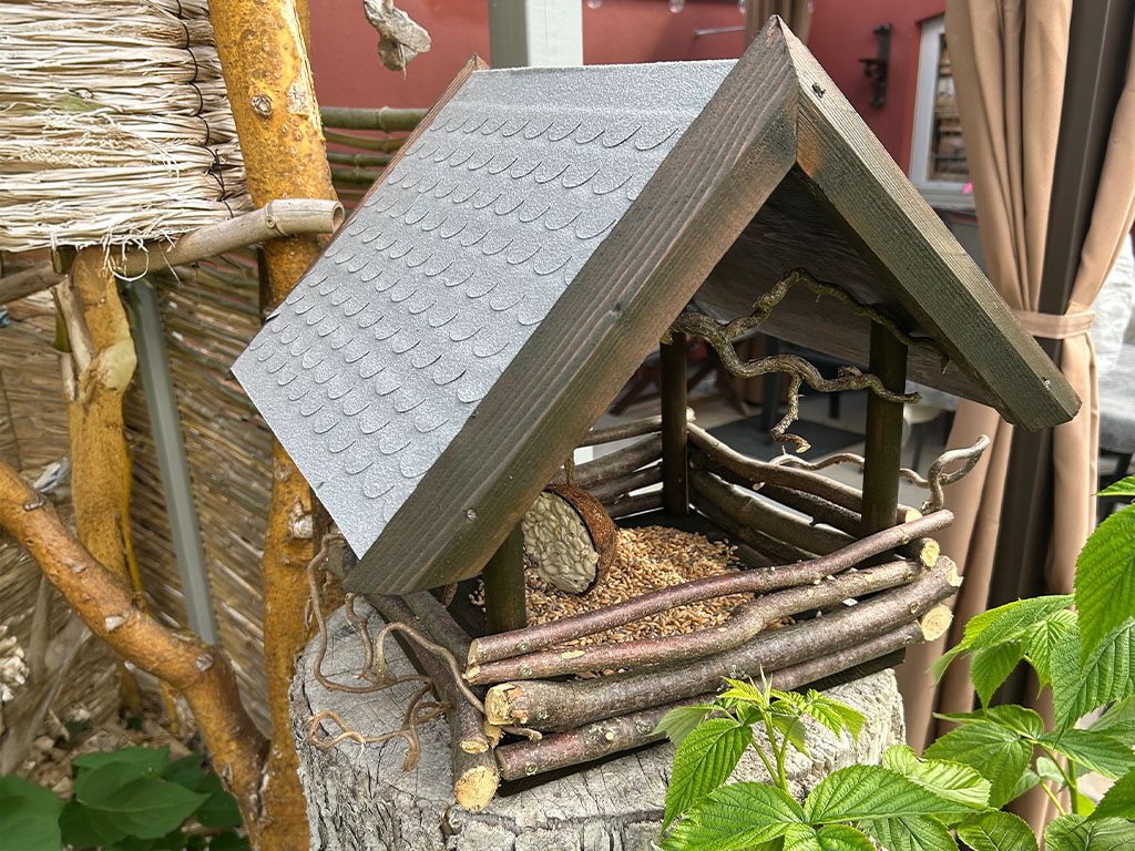Handmade bird feeder made of branches and wood with a grey roof, mounted on a tree stump with birdseed inside.