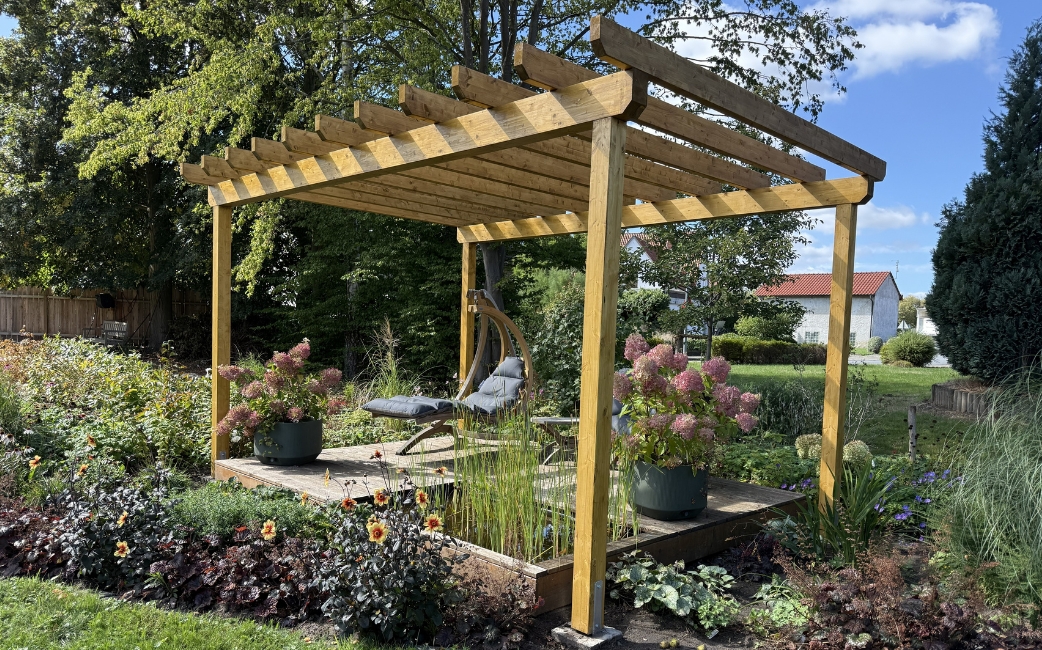Eine Pergola aus Holz mit einer Sitzecke, Hortensien in Töpfen und blühenden Pflanzen in einem Garten.