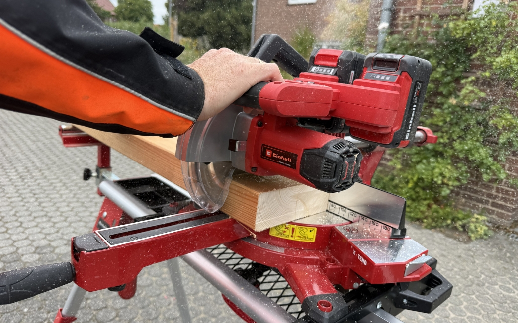 Nahaufnahme einer Person, die mit einer roten Einhell-Gehrungssäge einen Holzbalken zuschneidet.