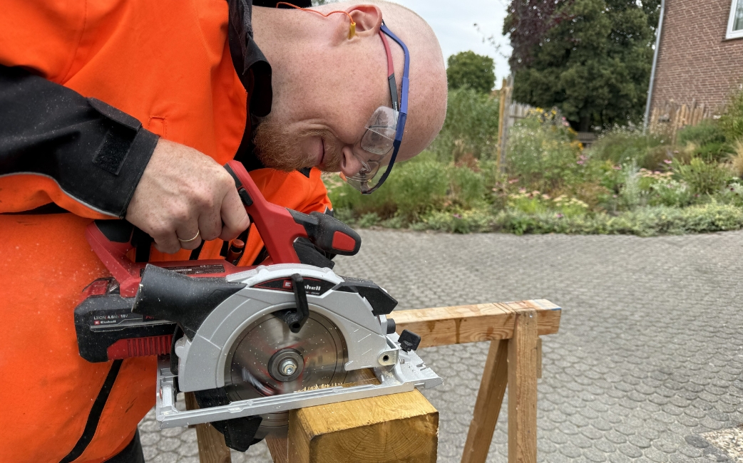 Eine Person, die eine Schutzbrille trägt, schneidet mit einer Handkreissäge ein Holzbrett, das im Freien auf einem Holzständer aufliegt.