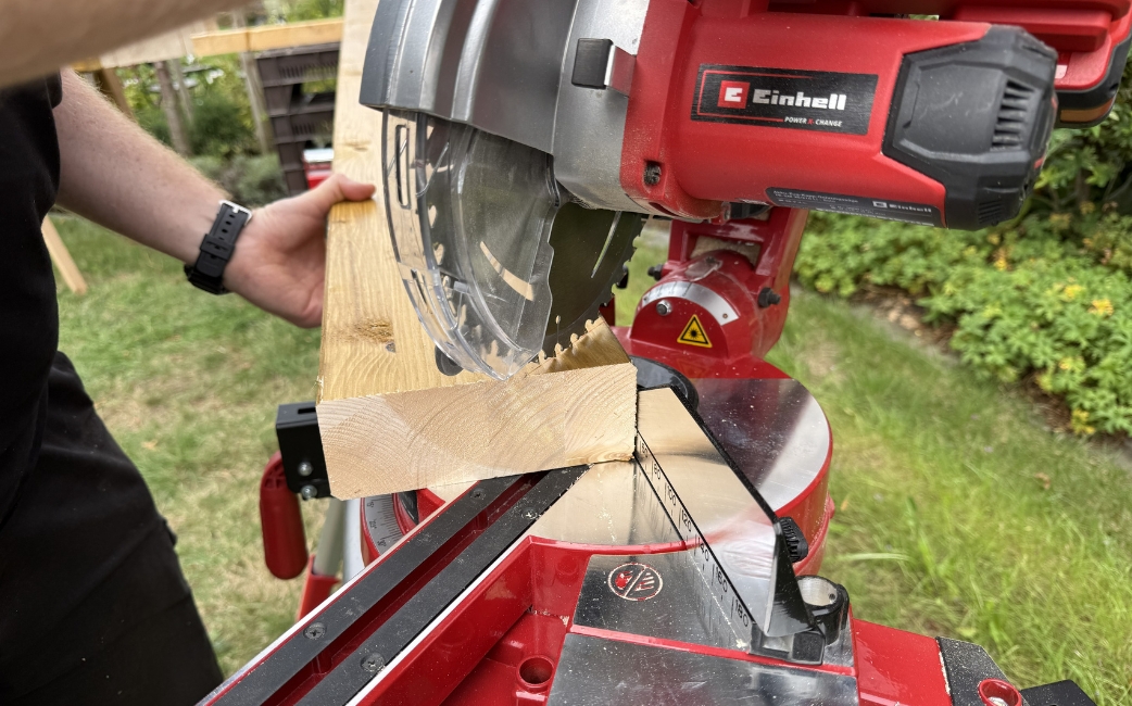 Nahaufnahme einer Einhell-Gehrungssäge beim Schneiden eines Holzbretts, wobei eine Hand das Werkstück festhält.