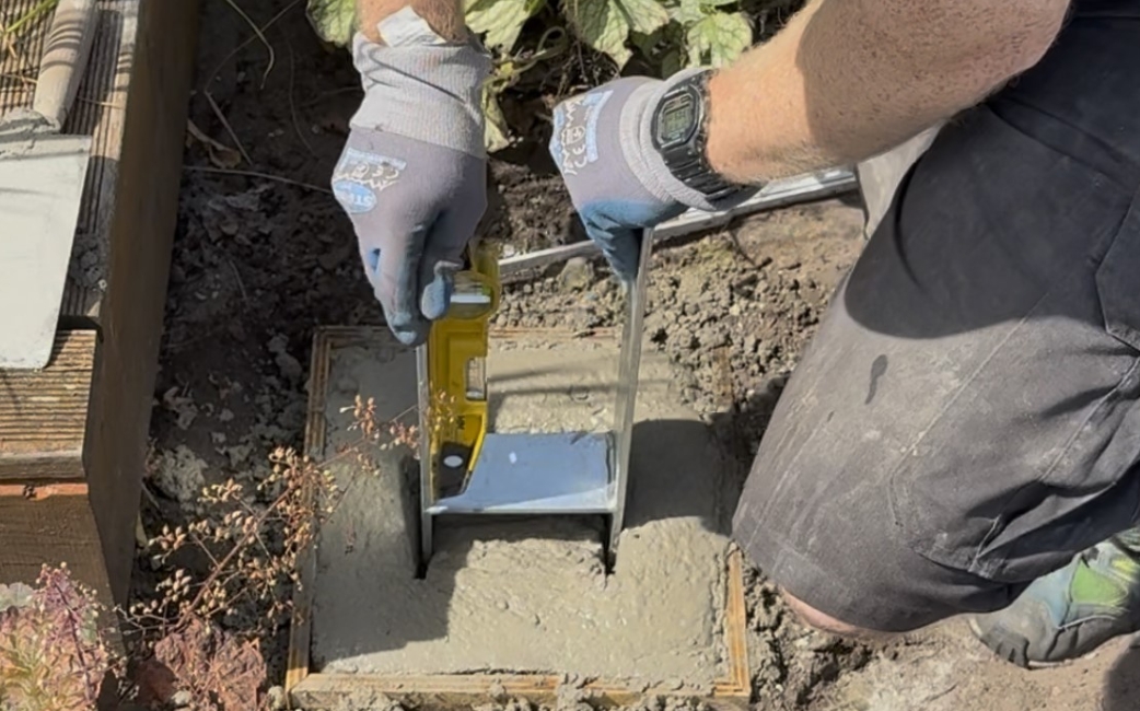 Eine Person, die Handschuhe trägt, bearbeitet mit einem gelben Handwerkzeug eine Metallhalterung, die in frischem Beton neben einem Holzbauwerk verankert ist.