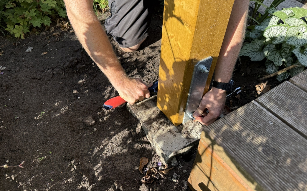 Eine Person befestigt einen Holzpfosten mit Hilfe einer Metallhalterung und eines Handwerkzeugs an einem Betonsockel.