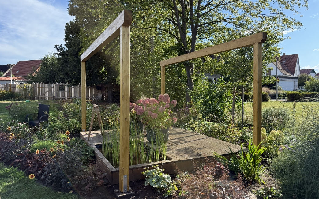 Fertiggestellter Pergola-Rahmen aus Holz auf einer erhöhten Gartenplattform, umgeben von Pflanzen.