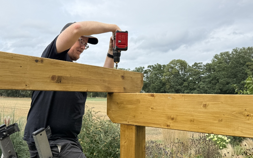  Eine Person befestigt im Freien mit einem Akku-Bohrschrauber einen Holzbalken am Rahmen einer Pergola.