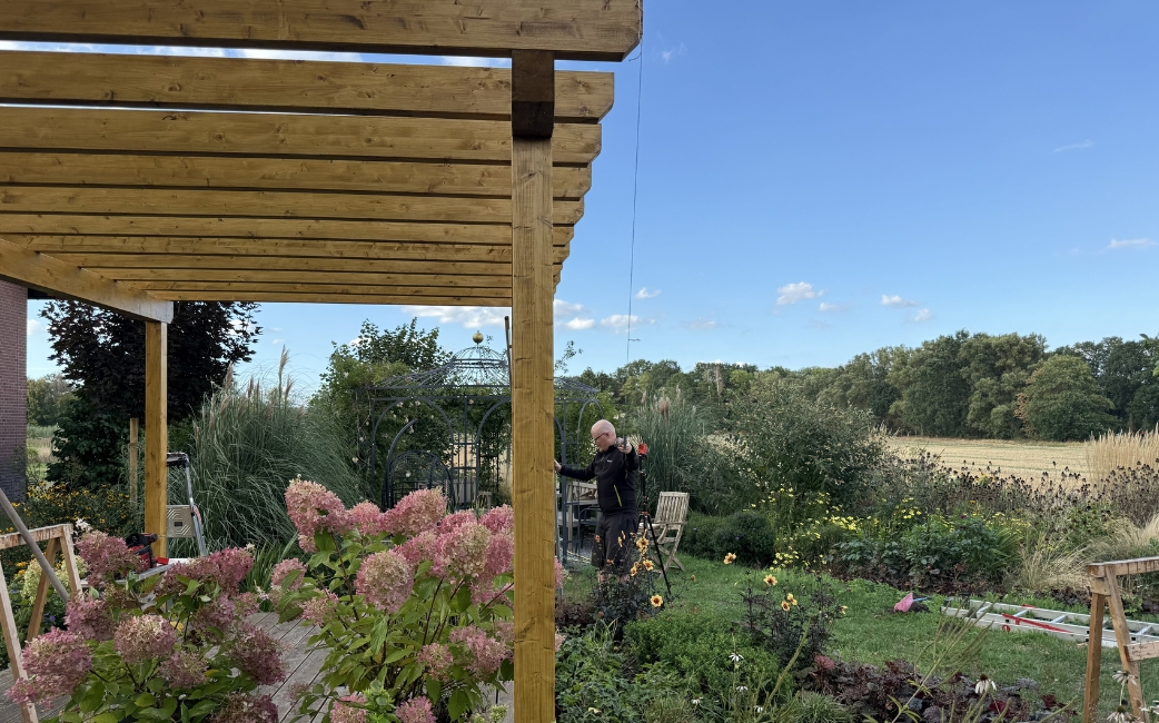 Fertiggestellte Holzpergola mit einem Lamellendach über einer Sitzecke im Garten, umgeben von blühenden Pflanzen und viel Grün.