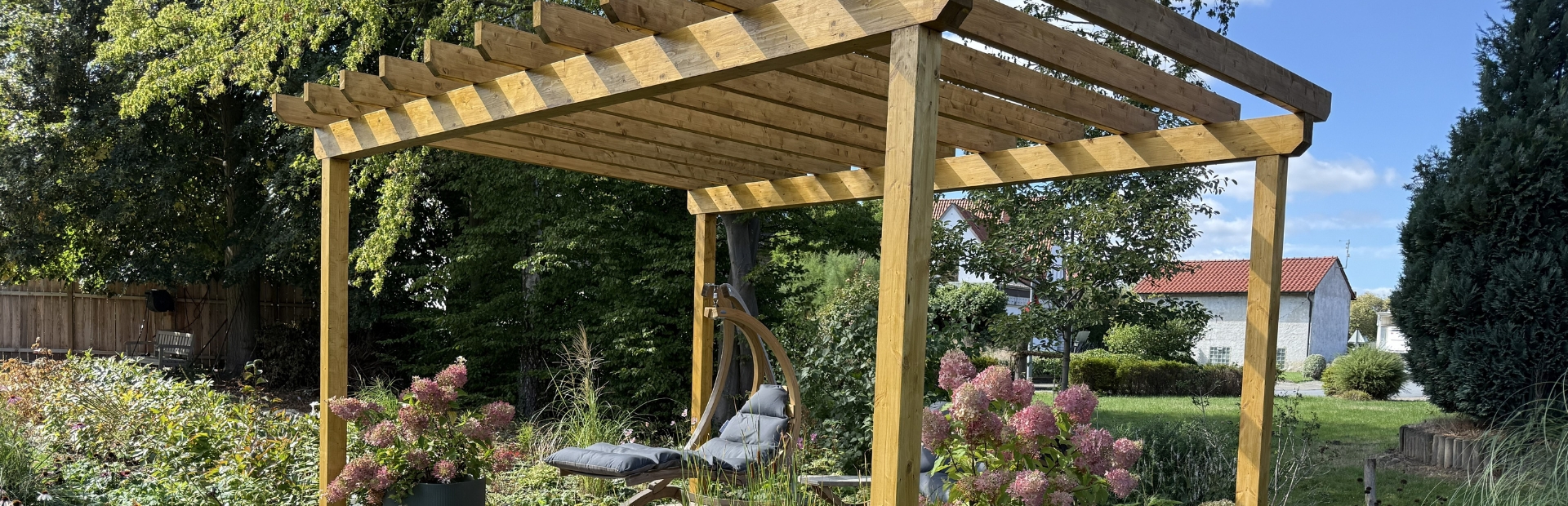 Eine Pergola aus Holz mit einer Sitzecke, Hortensien in Töpfen und blühenden Pflanzen in einem Garten.
