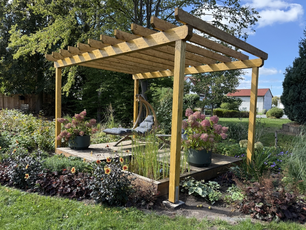Eine Pergola aus Holz mit einer Sitzecke, Hortensien in Töpfen und blühenden Pflanzen in einem Garten.