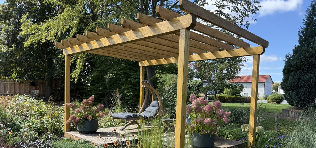 Eine Pergola aus Holz mit einer Sitzecke, Hortensien in Töpfen und blühenden Pflanzen in einem Garten.