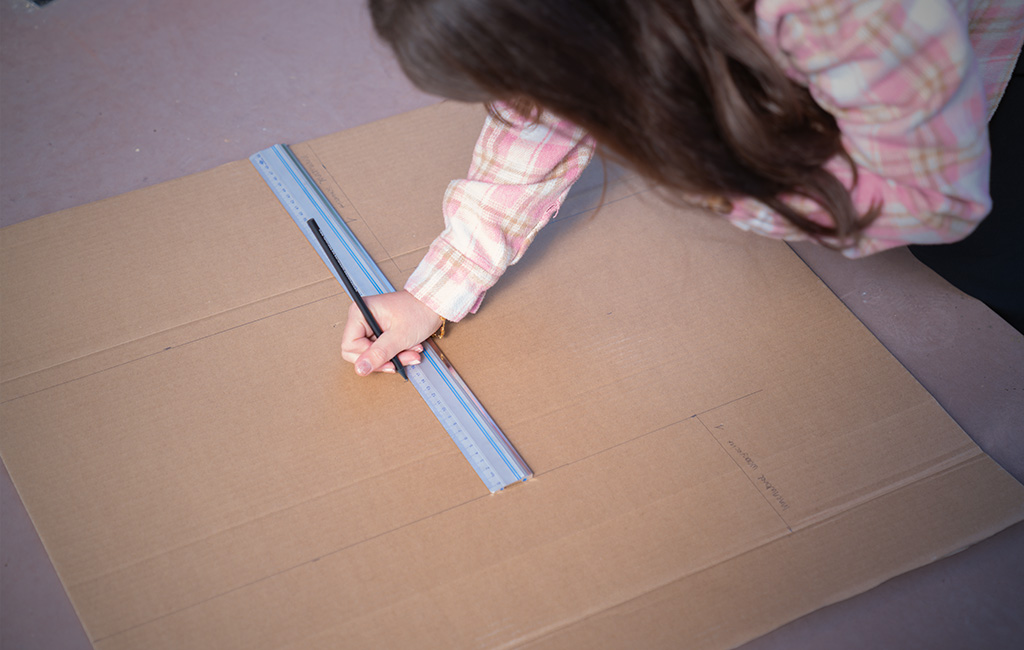 Person draws lines with a ruler on a large cardboard sheet.