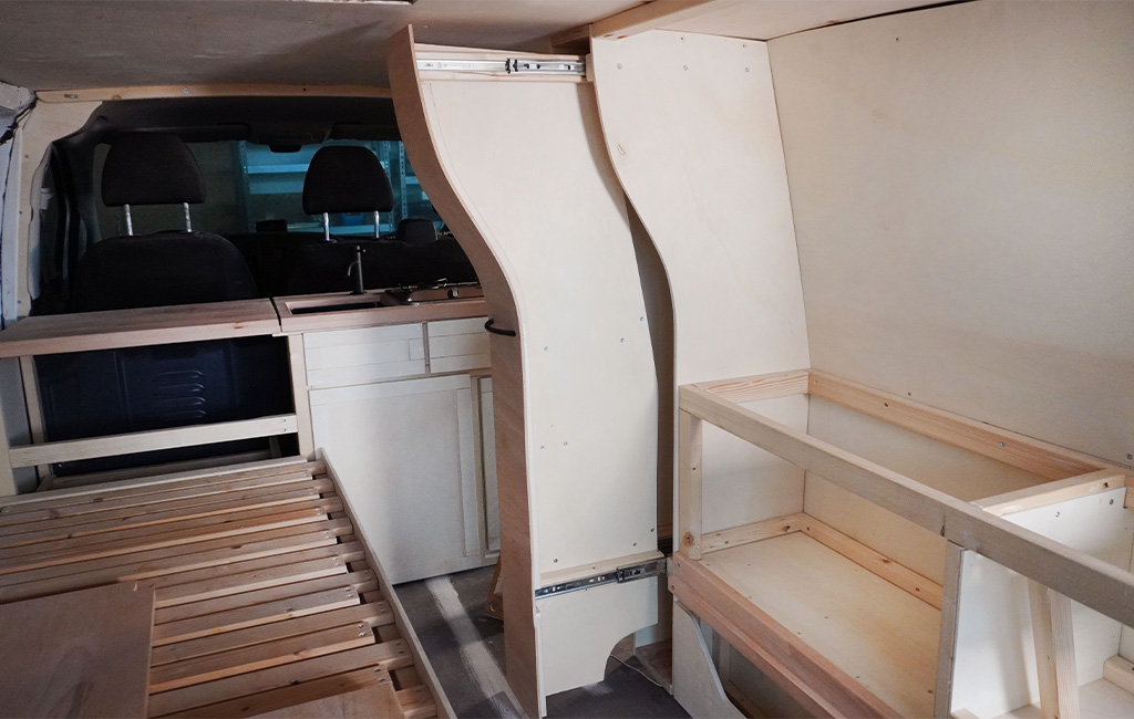 Interior view of the camper conversion with bed, kitchen, and closed bathroom cabinet.