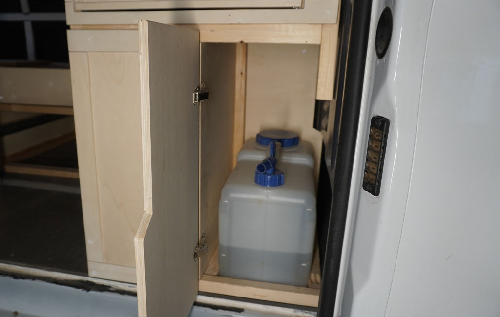 Two canisters with blue lids are placed behind an open wooden cabinet door in the camper.