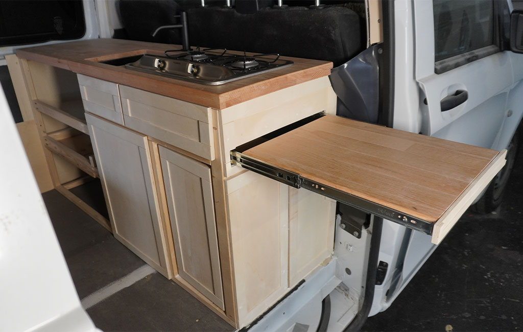 Fully installed camper kitchen with gas stove, sink, and pull-out side table on metal rails.