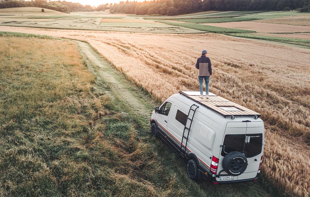 Camper-Van mit montierter Dachterrasse fährt auf einem Feldweg zwischen Feldern im Sonnenuntergang.