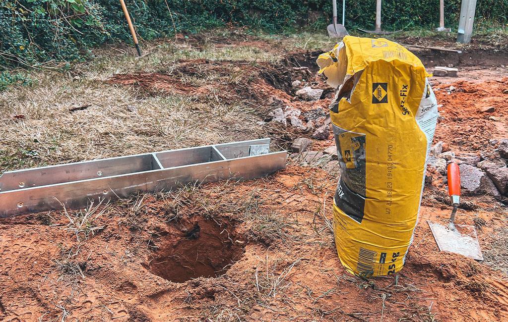 A torn yellow bag of concrete mix next to a hole in the ground and metal parts.