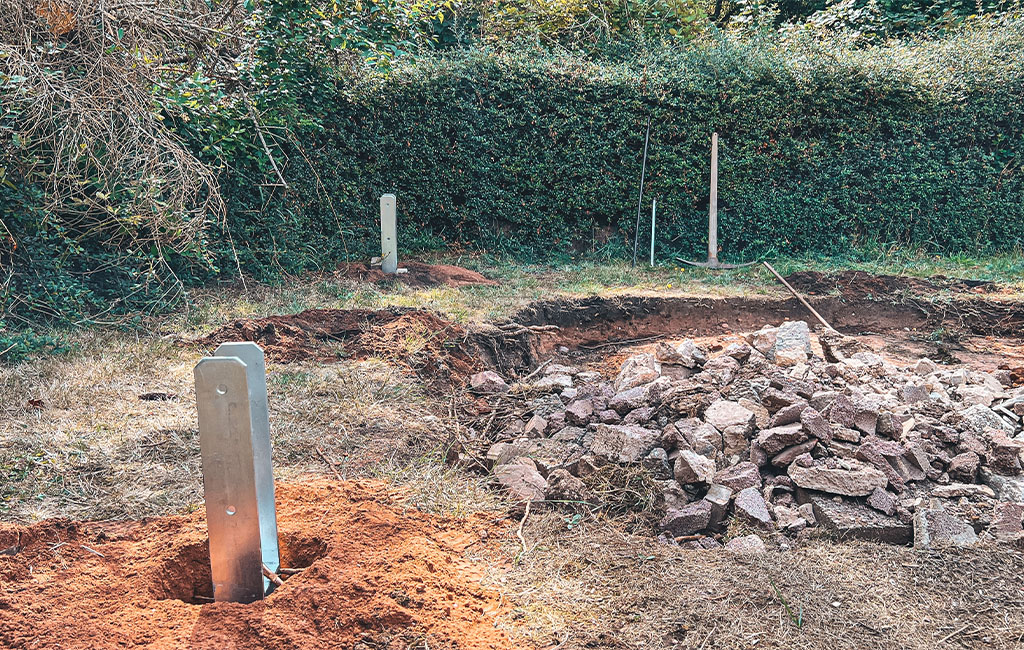 Several metal post bases placed in holes in the ground, with a pile of construction rubble nearby.