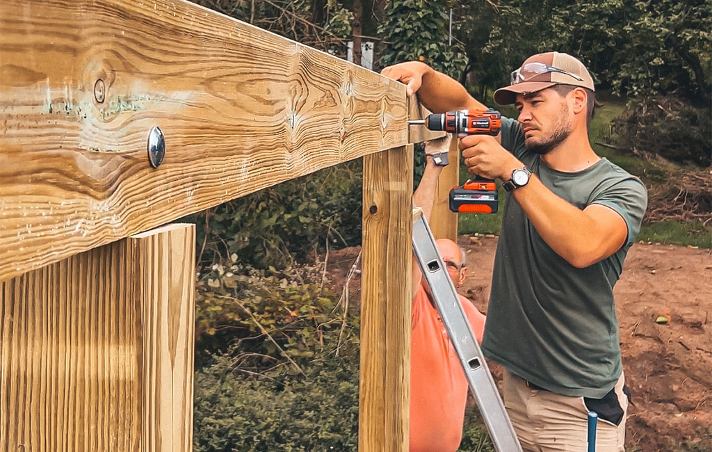 Two men assembling wooden beams using a cordless drill, one standing on a ladder.