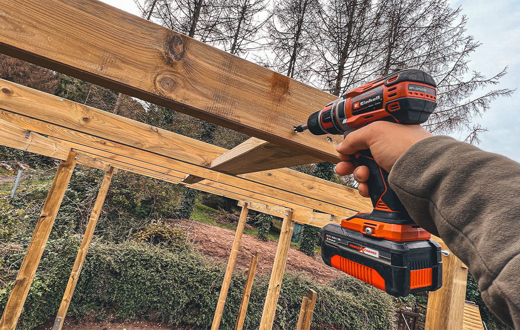 A person holds an Einhell cordless drill and screws together wooden beams for a carport.