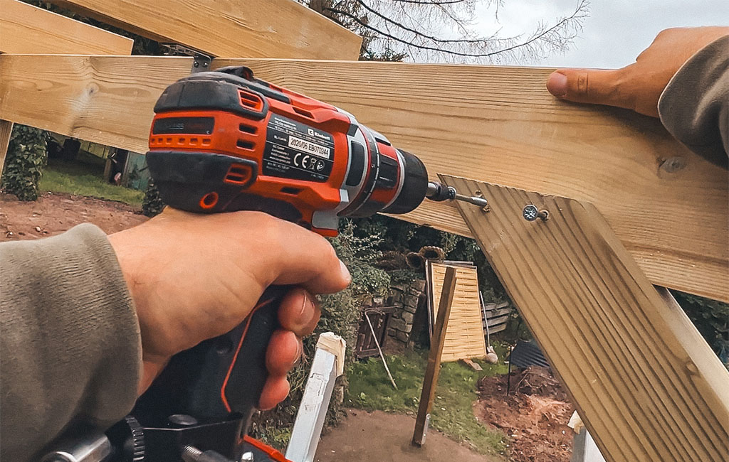 Close-up of a hand with an Einhell cordless drill screwing a wooden brace to a beam.