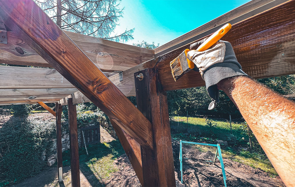 A person paints the wooden posts of the carport with brown wood stain.