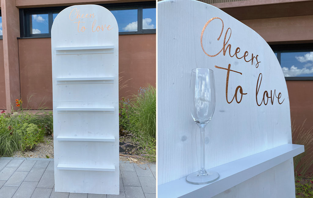 Zweiteilige Ansicht der fertig gestrichenen DIY-Sektwand mit fünf weißen Ablagen und goldener Aufschrift „Cheers to love“; rechts steht ein Sektglas auf einer Ablage.
