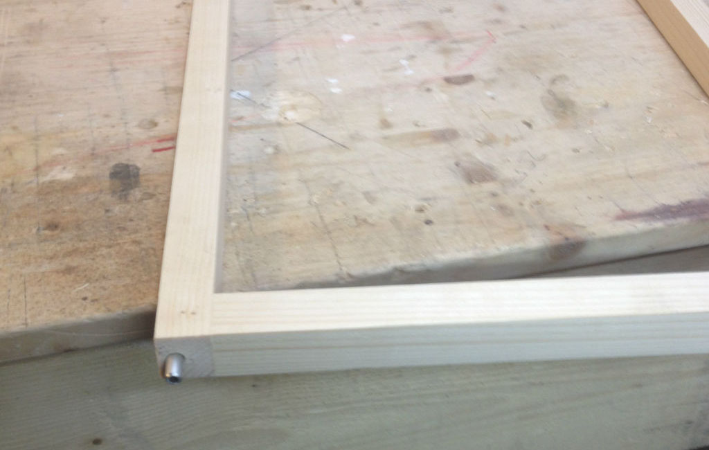 Nahaufnahme eines hellen Holzrahmens auf einer Werkbank. In einer Ecke ist ein Metallstift sichtbar.