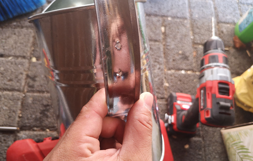 Eine Hand hält eine Metallgießkanne mit gebohrten Löchern, im Hintergrund ein Einhell Akkuschrauber.