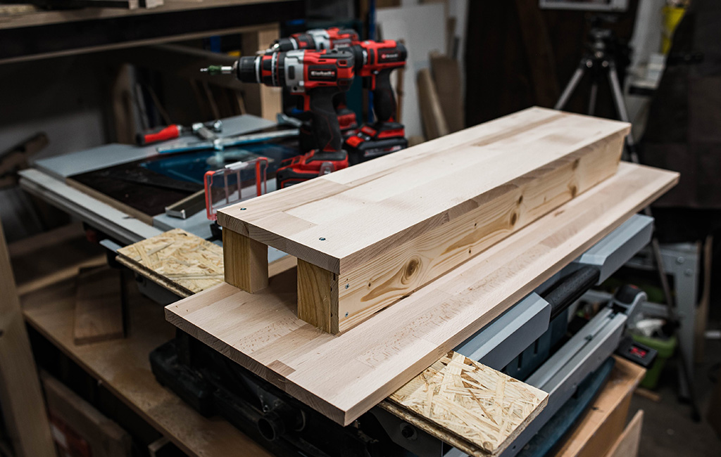 Partially assembled wooden structure on a workbench with Einhell cordless drill and tools in the background.