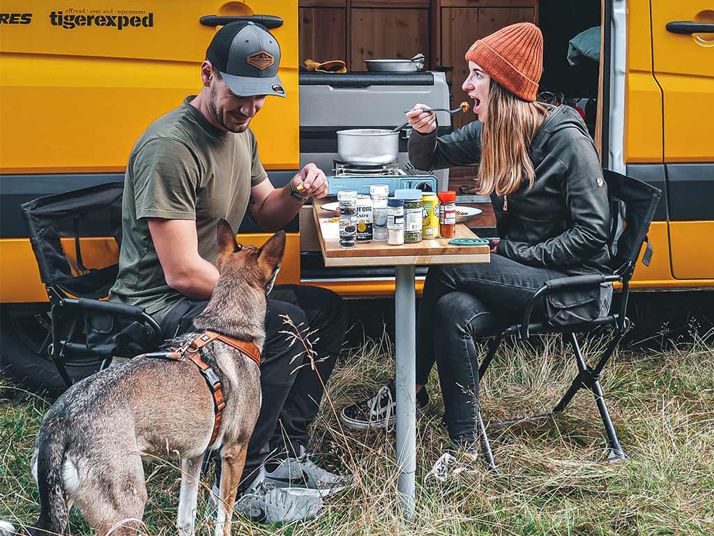 A couple sits at a folding table in front of an open camper, with a dog standing nearby.