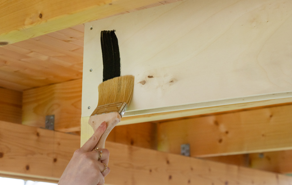 A hand applies black chalkboard paint to a light wooden board of the bar structure using a wide paintbrush.