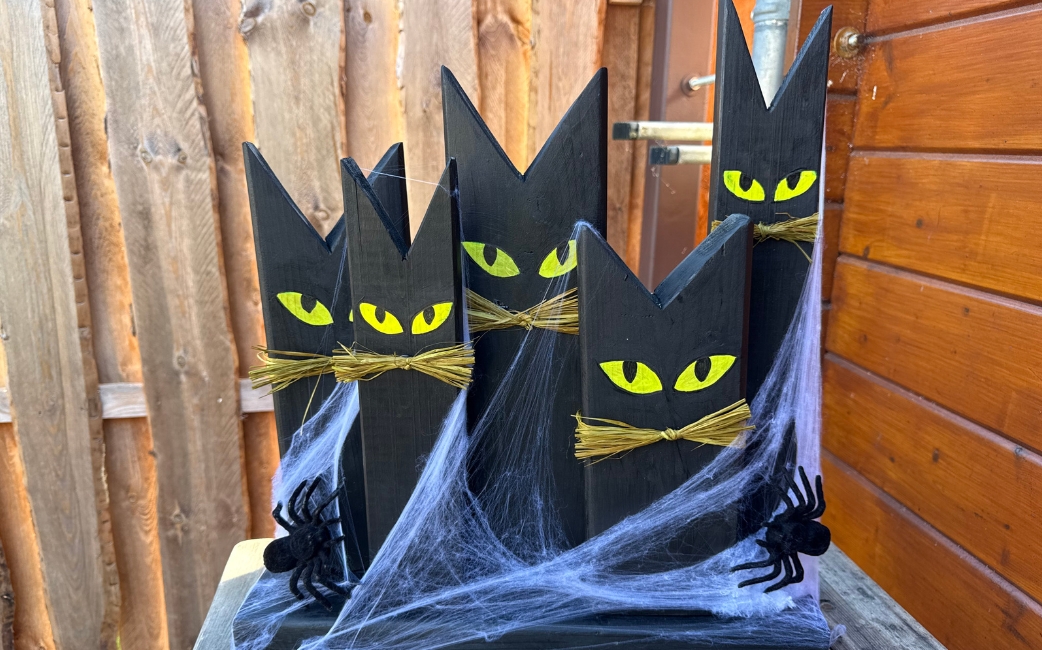 Fünf schwarze, katzenförmige Holzdekorationen mit gelben Augen und Strohhaaren stehen mit künstlichen Spinnweben und schwarzen Spinnen vor einem Holzzaun.