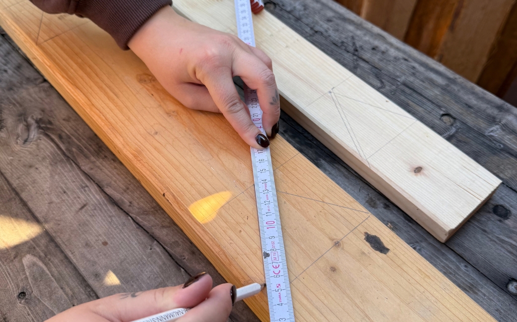 Eine Person misst ein Holzbrett mit einem Zollstock und markiert die Messstelle mit einem Bleistift.