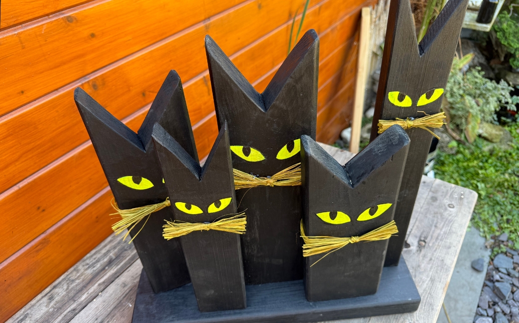 Fünf schwarze, katzenförmige Holzdekorationen mit gelben Augen und Schnurrhaaren aus Stroh stehen auf einem gemeinsamen Holzsockel.