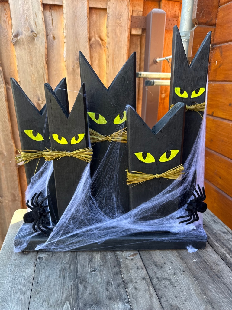 Fünf schwarze, katzenförmige Holzdekorationen mit gelben Augen und Schnurrhaaren aus Stroh stehen auf einem gemeinsamen Holzsockel.