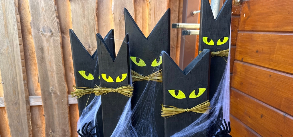 Fünf schwarze, katzenförmige Holzfiguren mit gelben Augen und Schnurrhaaren aus Stroh stehen auf einem gemeinsamen Holzsockel.