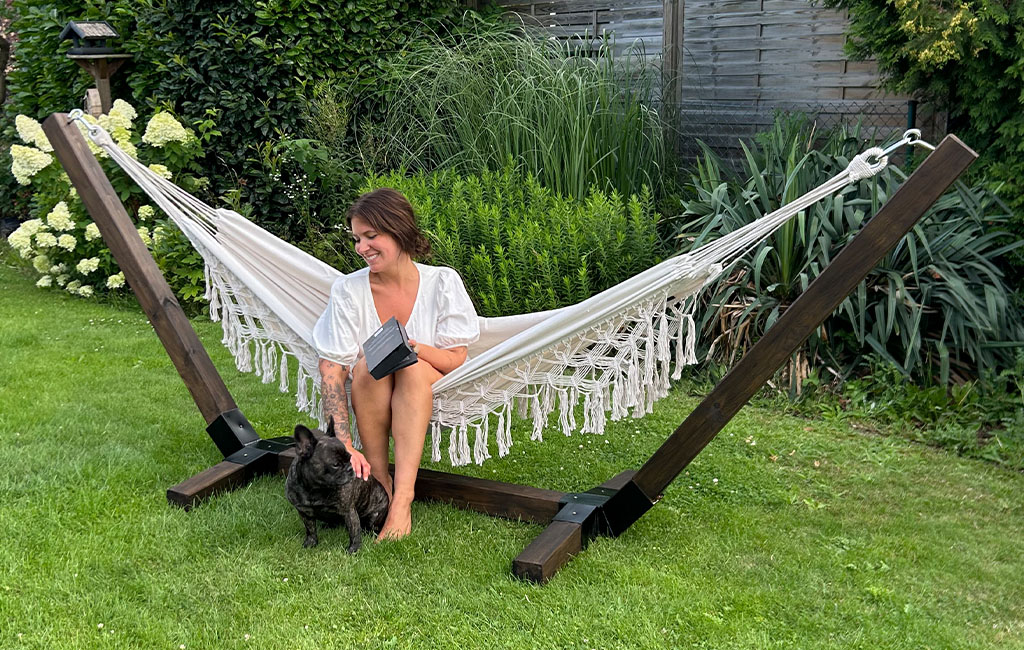 A woman sits in the hammock with a dark wooden frame in the garden and pets a black dog.