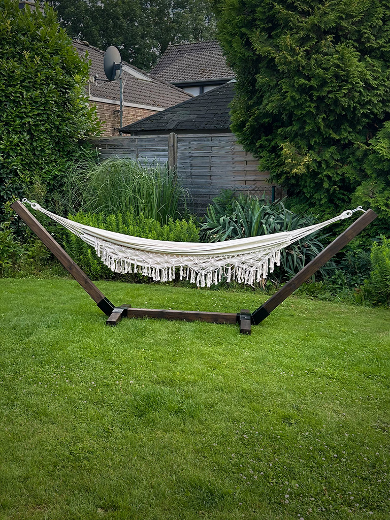 A cream-colored hammock with fringes hangs in a dark wooden frame on a lawn in a garden.