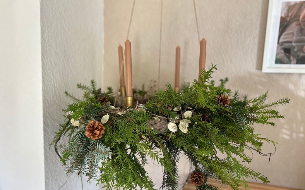 A hanging Advent wreath made of fir greenery with four brown taper candles, decorated with cones and dried plants.