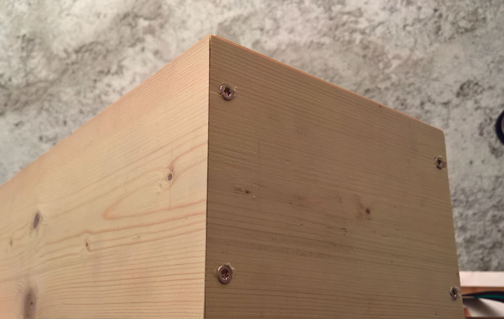 Nahaufnahme einer Ecke eines Holzkastens, verschraubt mit vier sichtbaren Schrauben.