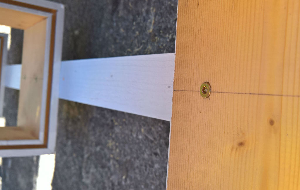 Ein senkrechtes, weiß lackiertes Holzbrett wird mit einer Schraube an einen Holzkasten geschraubt.