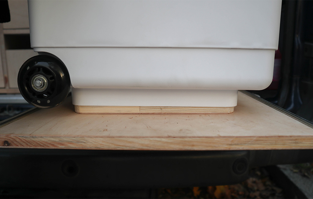 Weißer Kanister mit Rollen steht passgenau auf einer Halterung aus Holz im Heck eines Vans.